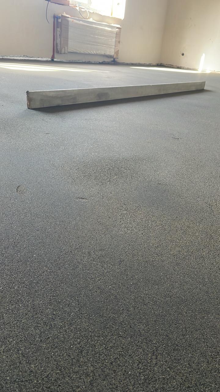 Крупный план свежей тёмно-серой стяжки с правилом и радиатором на заднем плане.